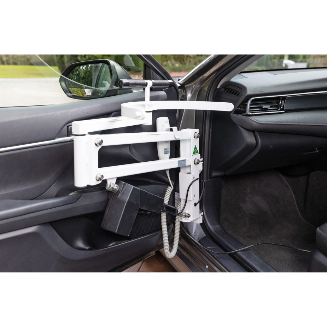 A white Para Mobility IBIS Wheelchair to Car Access Lift is mounted inside the car door near the driver’s seat. It has metal arms, a black control box, and a flexible tube for smooth wheelchair-to-car transfers. The car door is open.